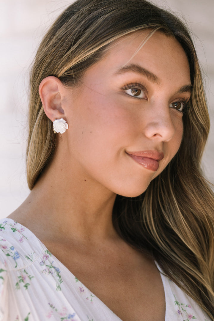 A close-up portrait of a woman with long, wavy brown hair and soft makeup, wearing small, white flower-shaped earrings.