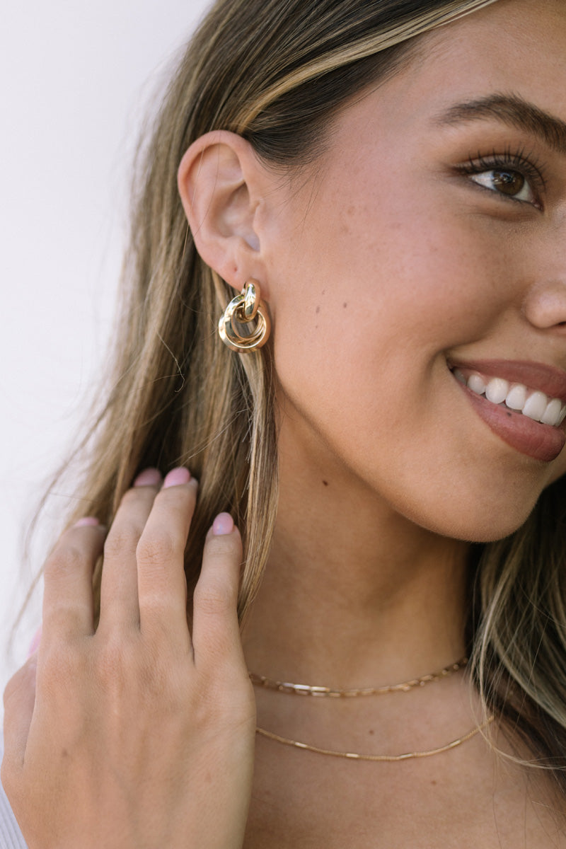 Close-up image of a woman's face, smiling and wearing gold knot dangle earrings with a necklace.