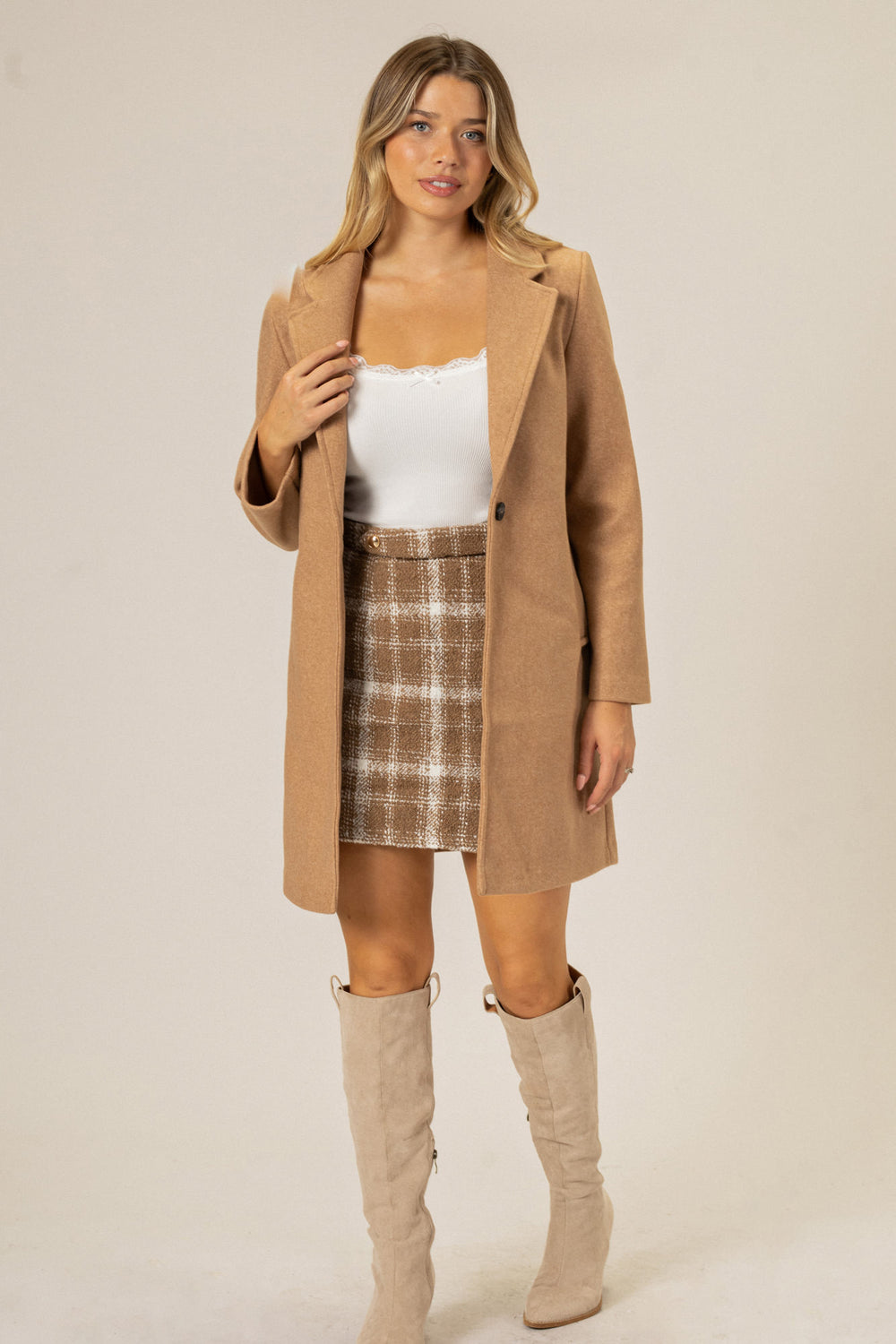 A beige notched-lapel coat worn over a white top and a plaid skirt, paired with tall beige boots.