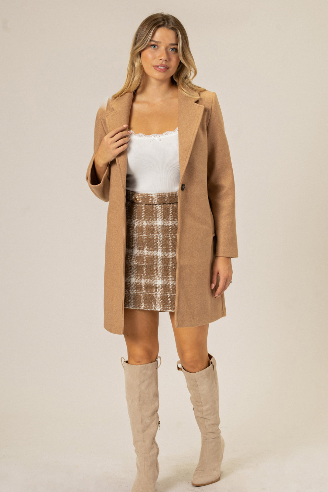 A beige notched-lapel coat worn over a white top and a plaid skirt, paired with tall beige boots.