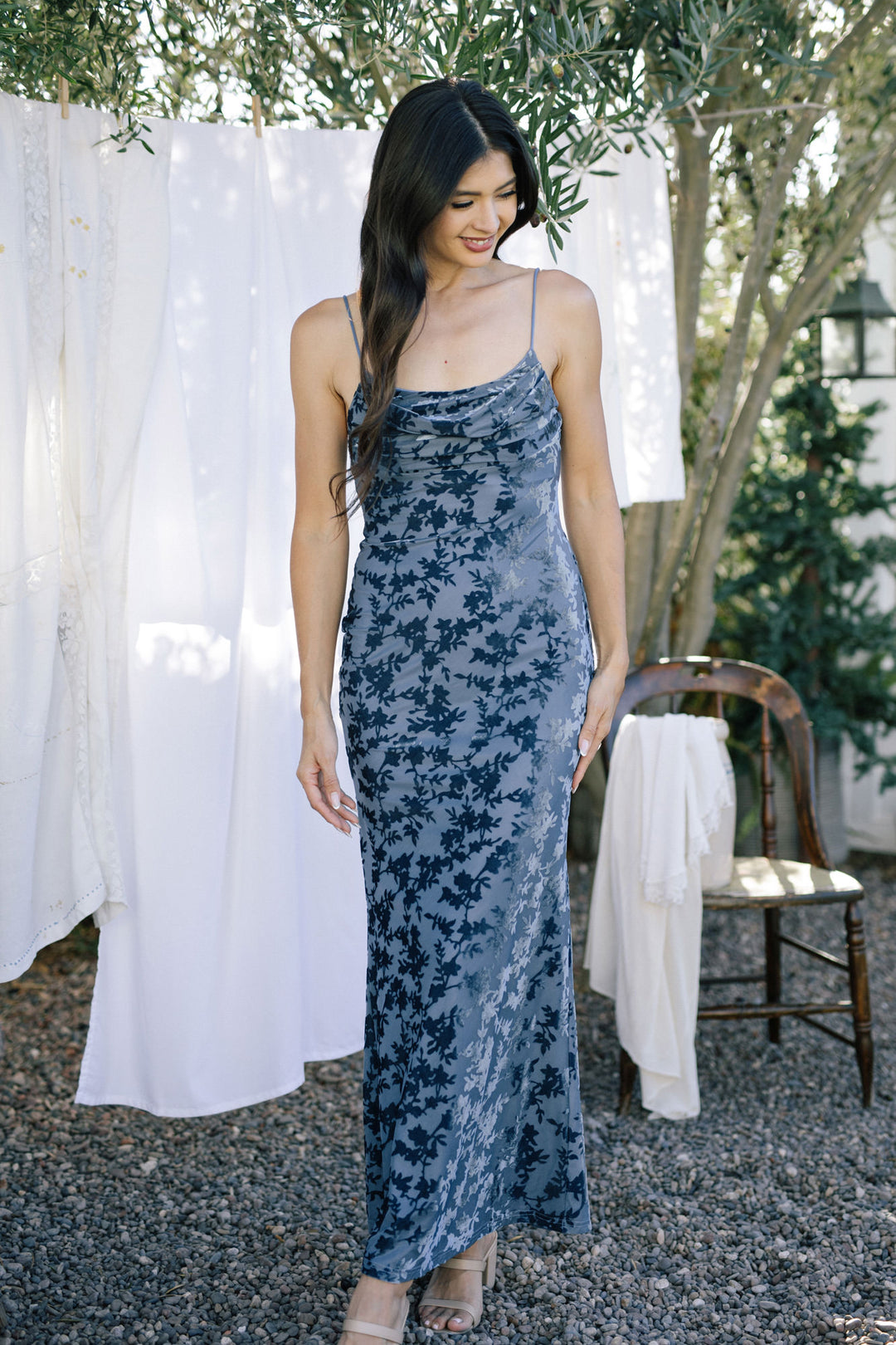 A model wearing a blue floral velvet dress with spaghetti straps, a fitted bodice, and a flowing maxi skirt, posing in a garden setting.