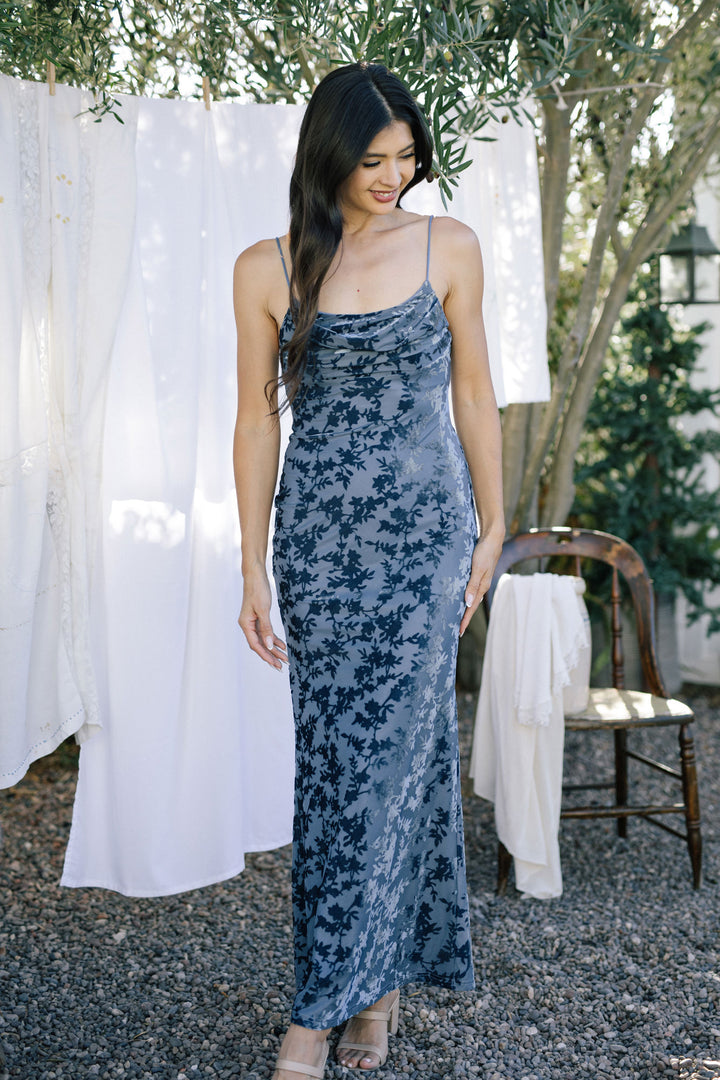 A model wearing a blue floral velvet dress with spaghetti straps, a fitted bodice, and a flowing maxi skirt, posing in a garden setting.