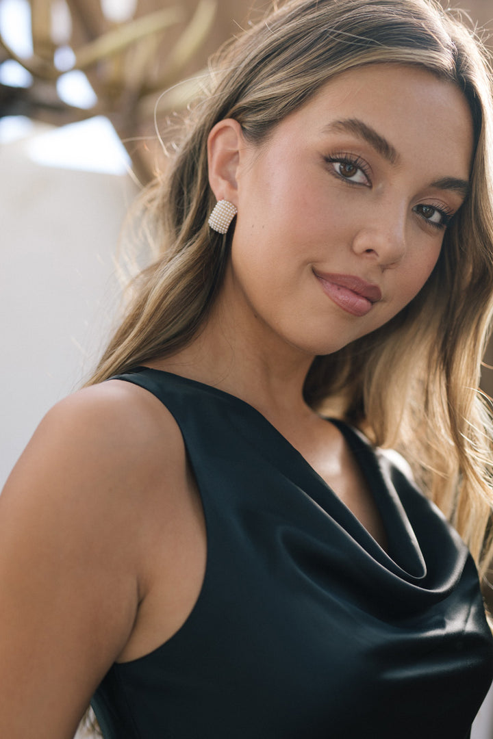 A woman with long blonde hair and pearl stud earrings wears a black sleeveless dress, looking directly at the camera with a slight smile.