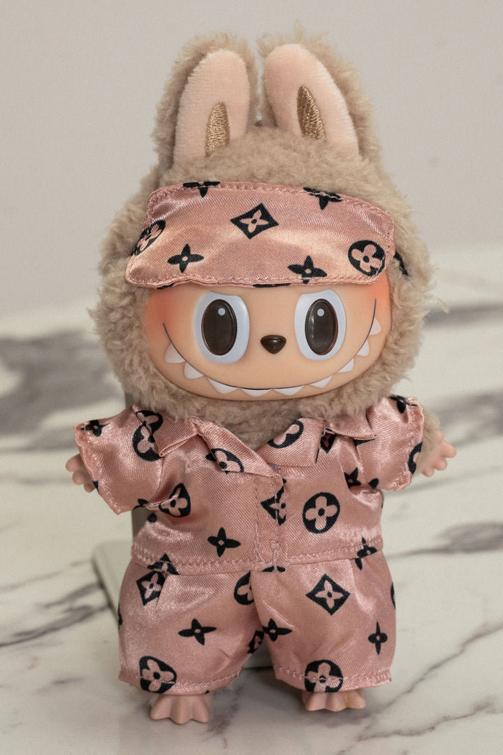 Beige plush stuffed animal wearing pink satin pajamas with black floral pattern, gold glitter accents, and a matching sleep mask.