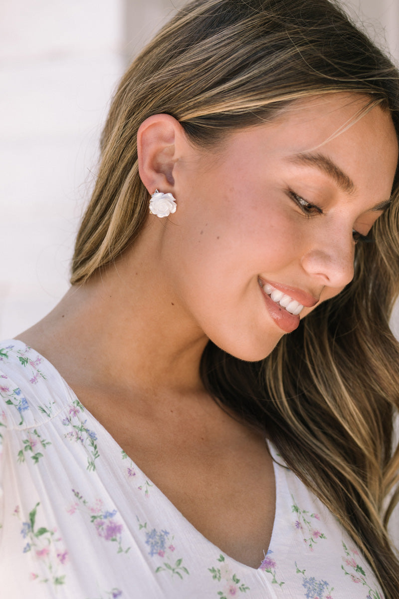 A close-up of a woman's face, smiling and wearing small white flower-shaped earrings against a soft floral backdrop.