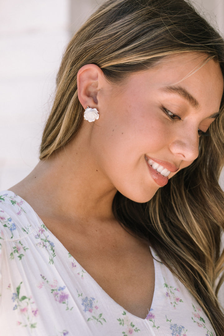 A close-up of a woman's face, smiling and wearing small white flower-shaped earrings against a soft floral backdrop.