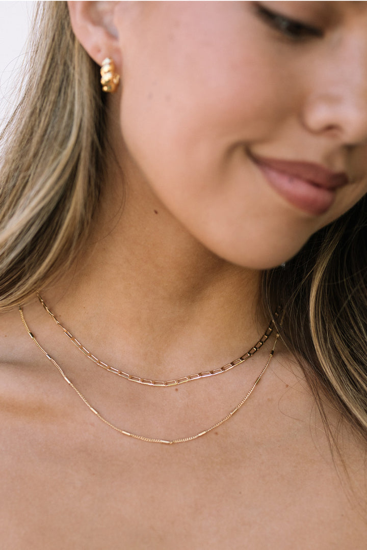 A close-up image of a woman's neckline, showcasing a layered gold necklace with a chain-link design.
