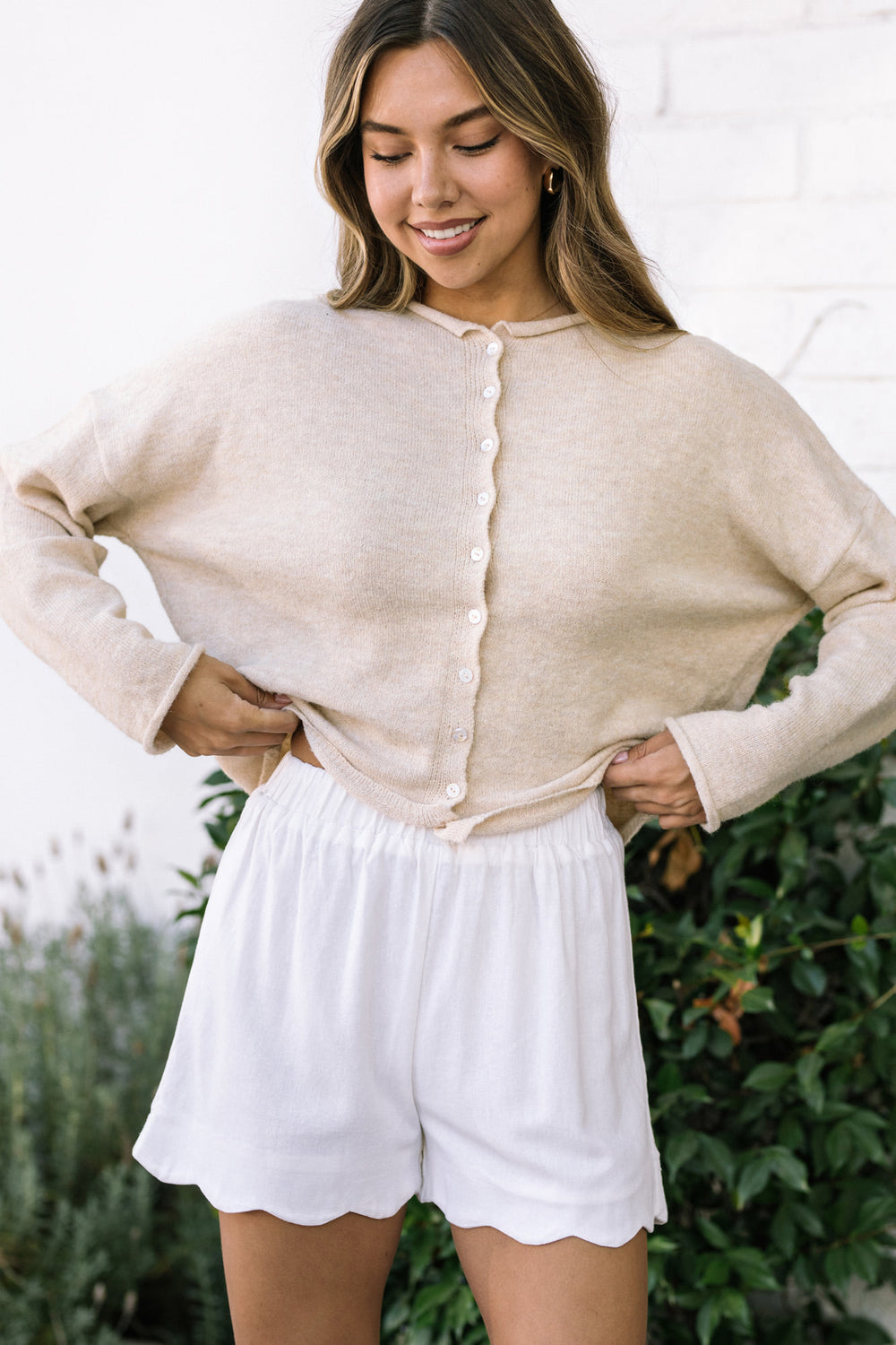 A cozy, ivory-colored cardigan with a relaxed, oversized fit and button closure, paired with white, high-waisted shorts with a scalloped hem.