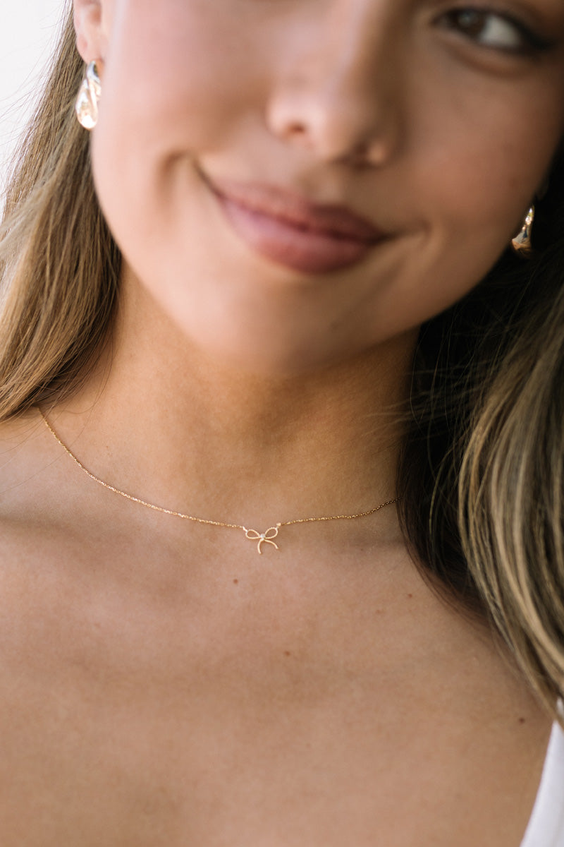 Close-up image of a delicate gold bow necklace with a thin chain, worn by a woman with long, wavy blonde hair.