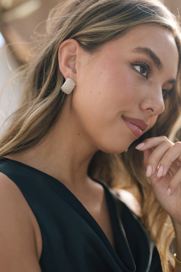Elegant pearl stud earrings with a sparkling crystal-encrusted face, framing a young woman's soft facial features and wavy blonde hair.