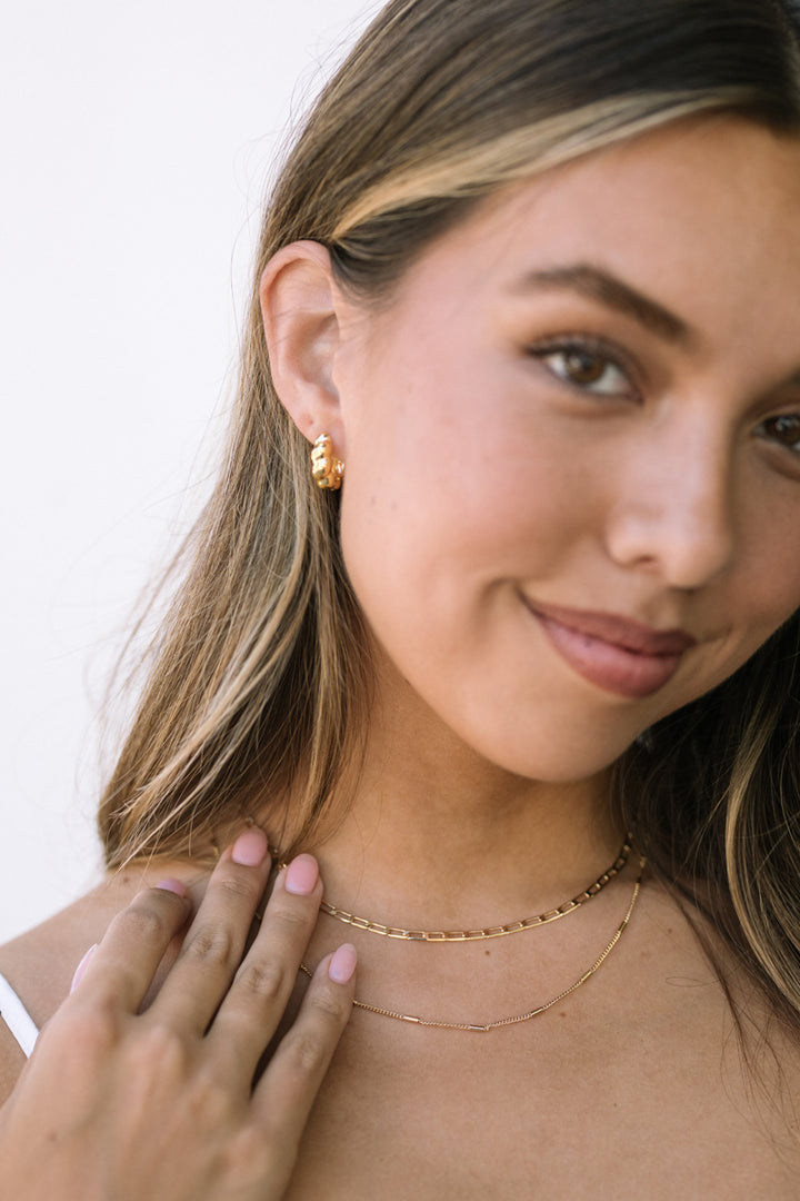 A close-up view of a woman's face, with a warm smile and layered gold necklaces and earrings adorning her.