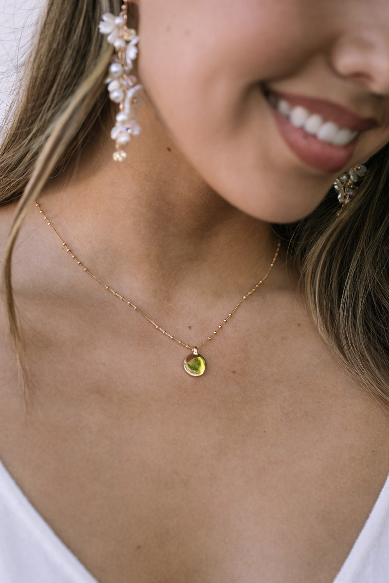 A close-up view of a woman's face, smiling and wearing a delicate gold-toned necklace with a green pendant.