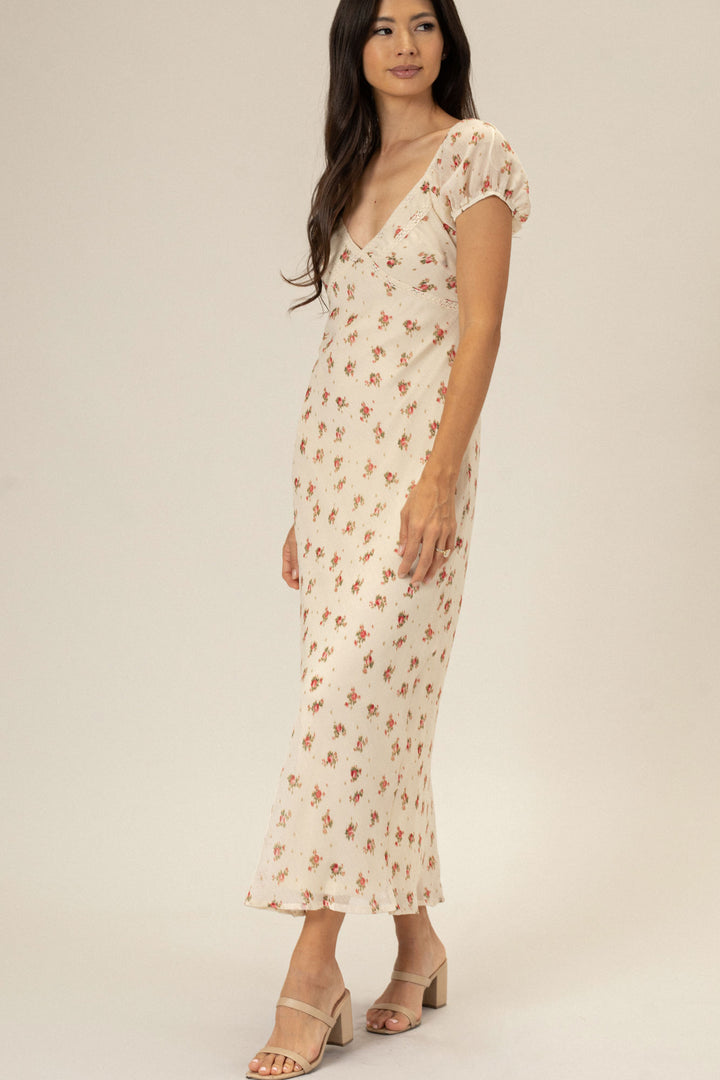 A woman wearing a floral print V-neck dress with puff sleeves, flowing to a maxi length, posing against a neutral background.