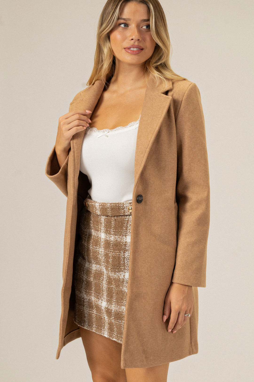 A camel-colored, single-breasted coat with notched lapels, worn over a white lace-trimmed tank top and patterned mini skirt, creating a stylish, layered look.
