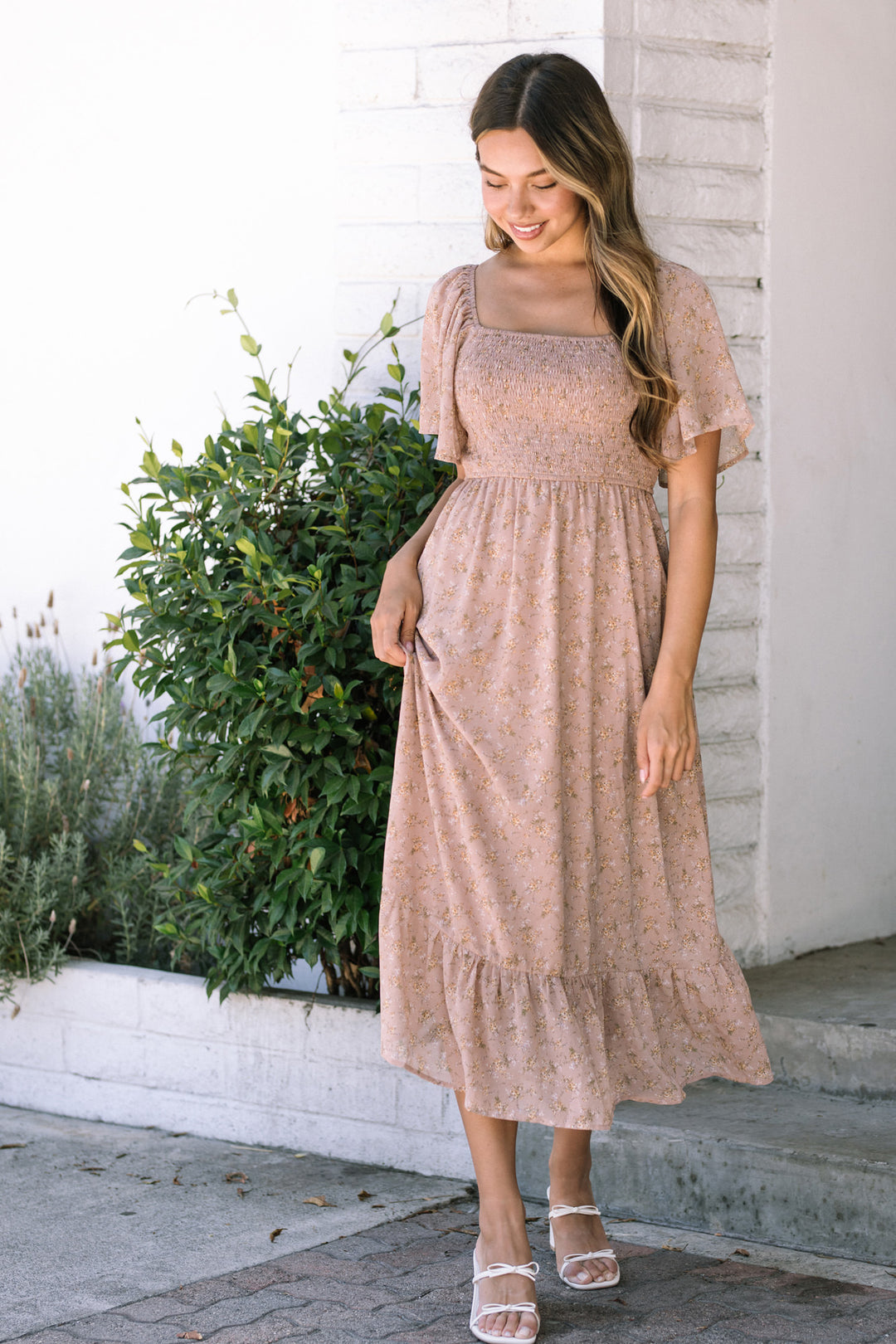A smocked midi dress in a light pink floral print, with short flutter sleeves and a tiered skirt, worn by a smiling woman with long wavy hair standing in front of greenery.