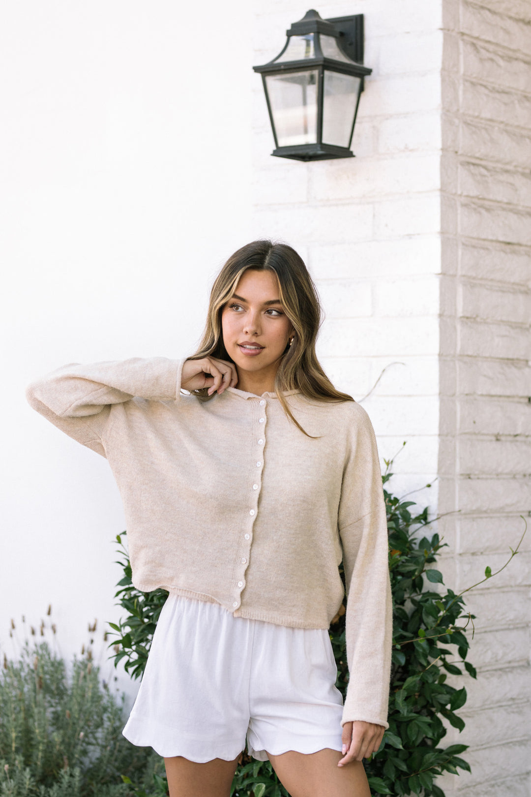 A woman wearing a light beige cardigan and white shorts, posing in front of a brick wall with a lantern above.