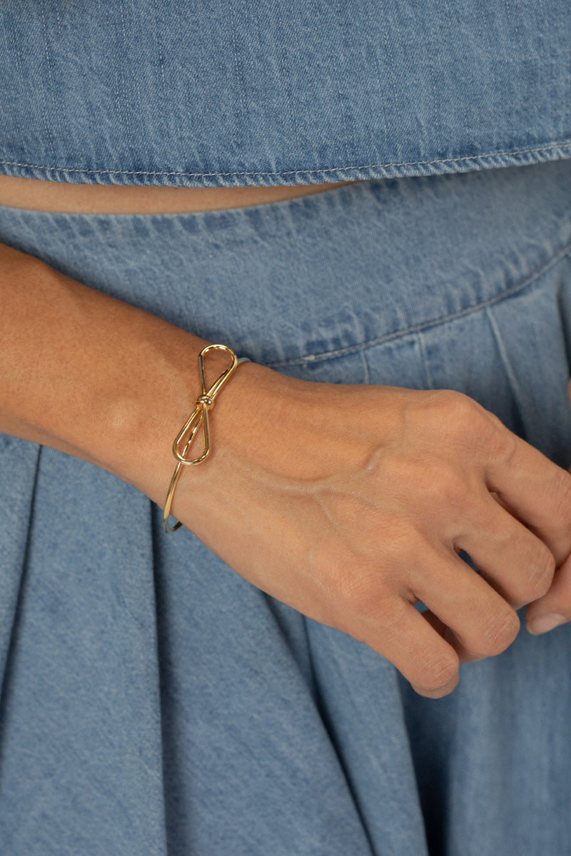 Gold-toned wire bow bracelet on a person's wrist, against a denim backdrop.