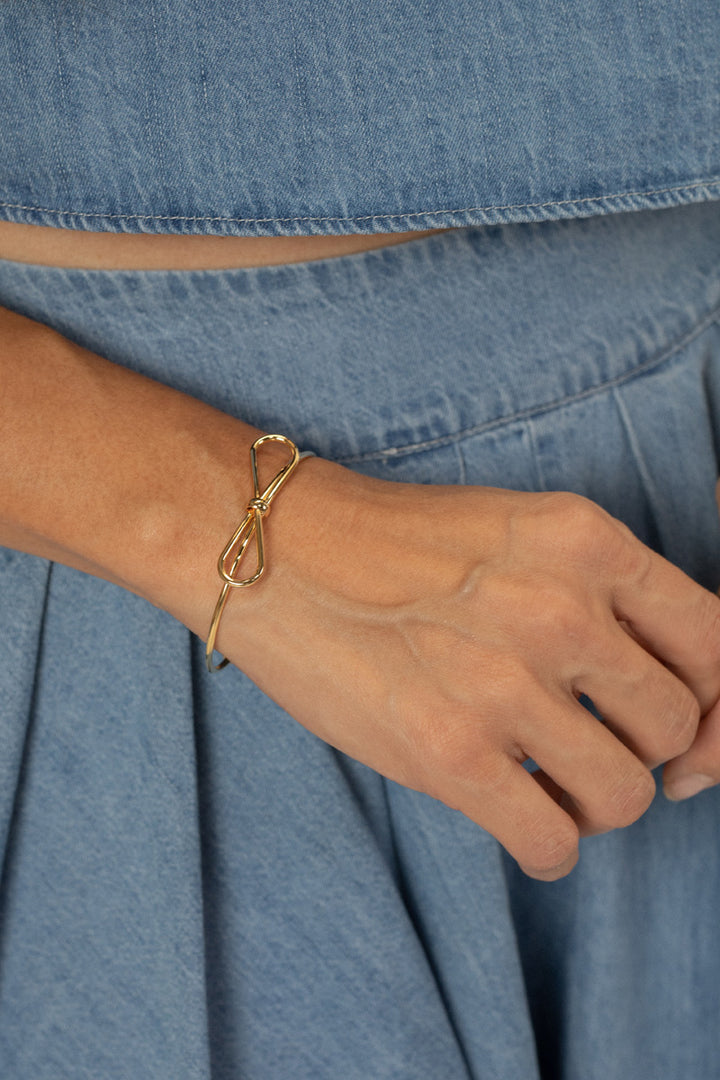 Gold-toned wire bow bracelet on a person's wrist, against a denim backdrop.