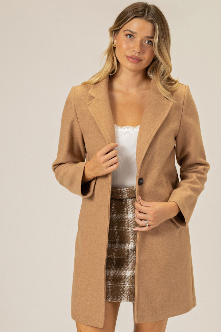 A camel-colored, notched lapel coat with a single button closure, worn over a white ribbed tank top and a printed skirt by a young woman with blonde hair.