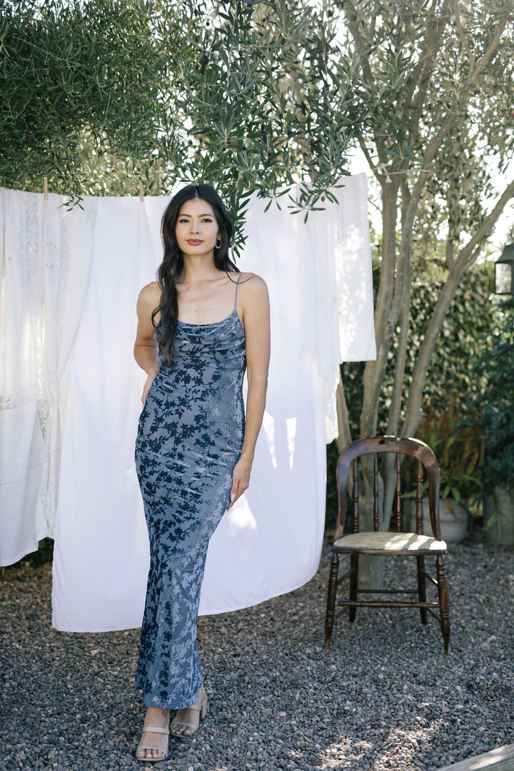 A woman wearing a blue, tie-dye patterned velvet maxi dress, posing in front of a white backdrop with greenery in the background.