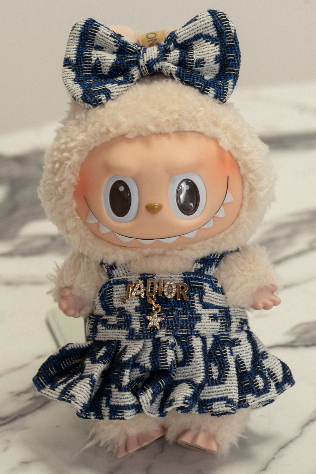 Plush Labubu doll in blue and white checkered dress, with a bow headband and "J'Dior" logo charm.
