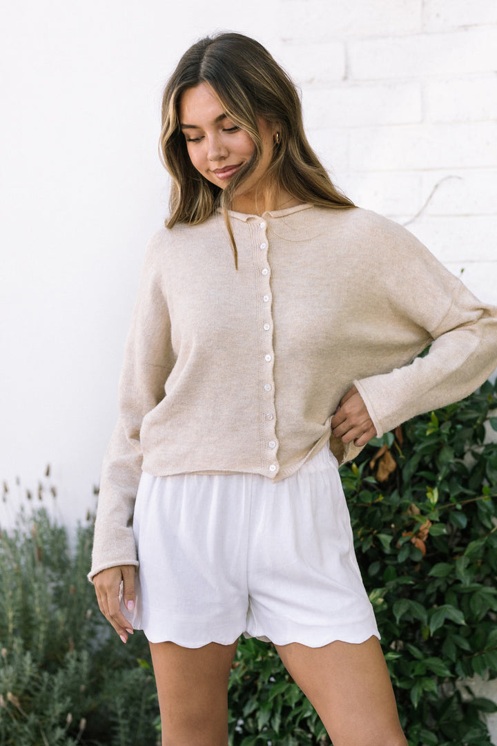 A pair of soft white scalloped shorts with a high elastic waistband, shown on a model with long brown hair standing in a garden.
