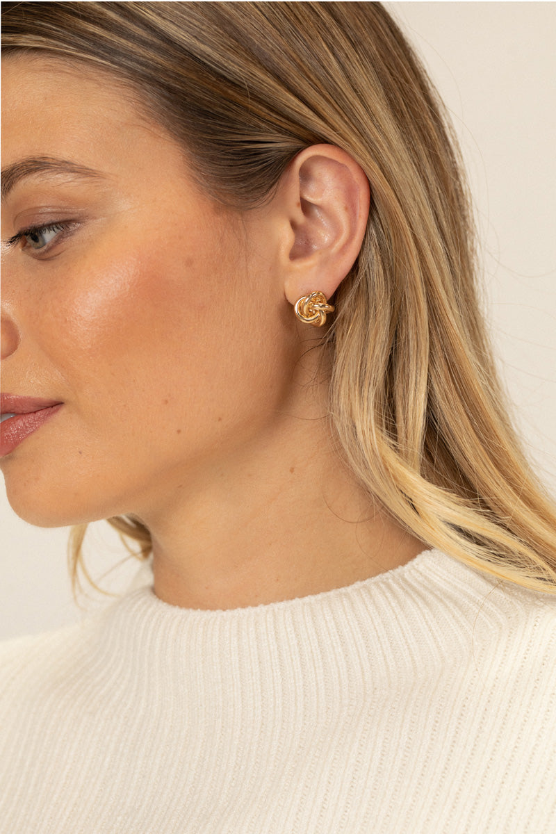 A close-up side view of a woman's ear, featuring a pair of gold-toned knotted stud earrings.