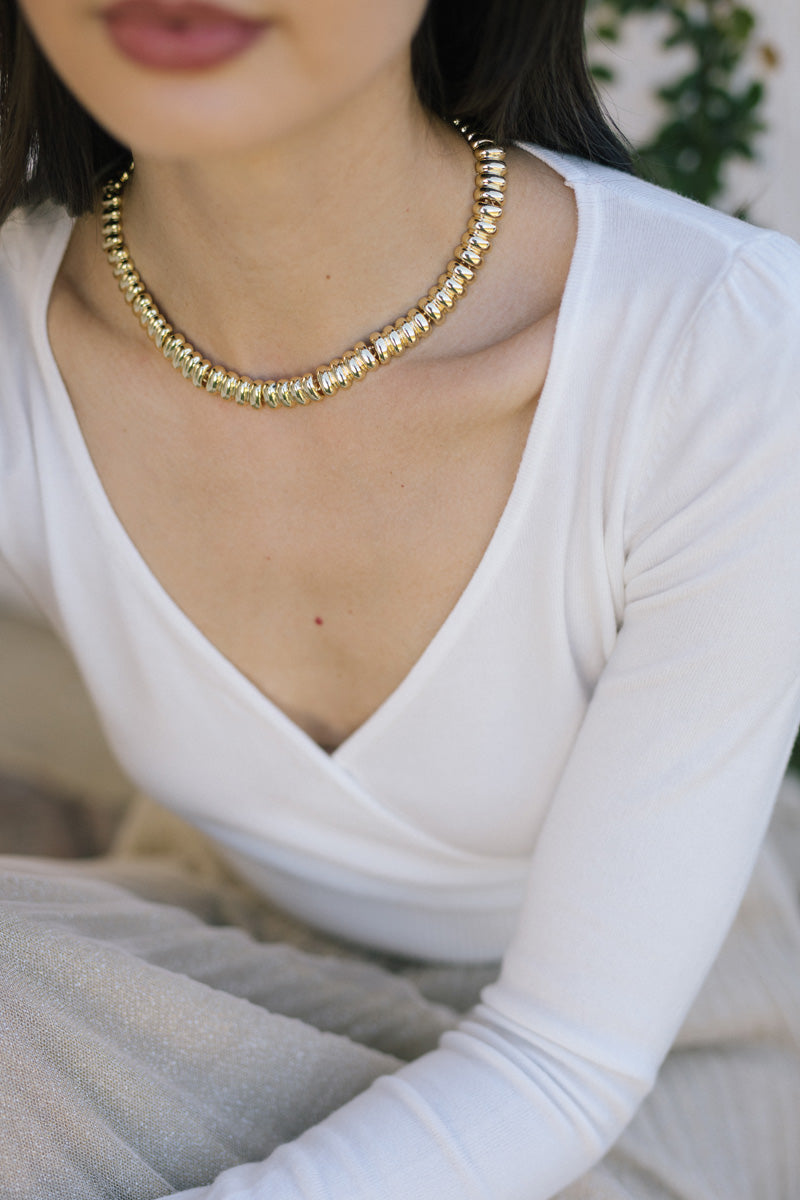 A close-up image of a woman wearing a gold-toned, beaded necklace featuring a repeating pattern of cylindrical beads.