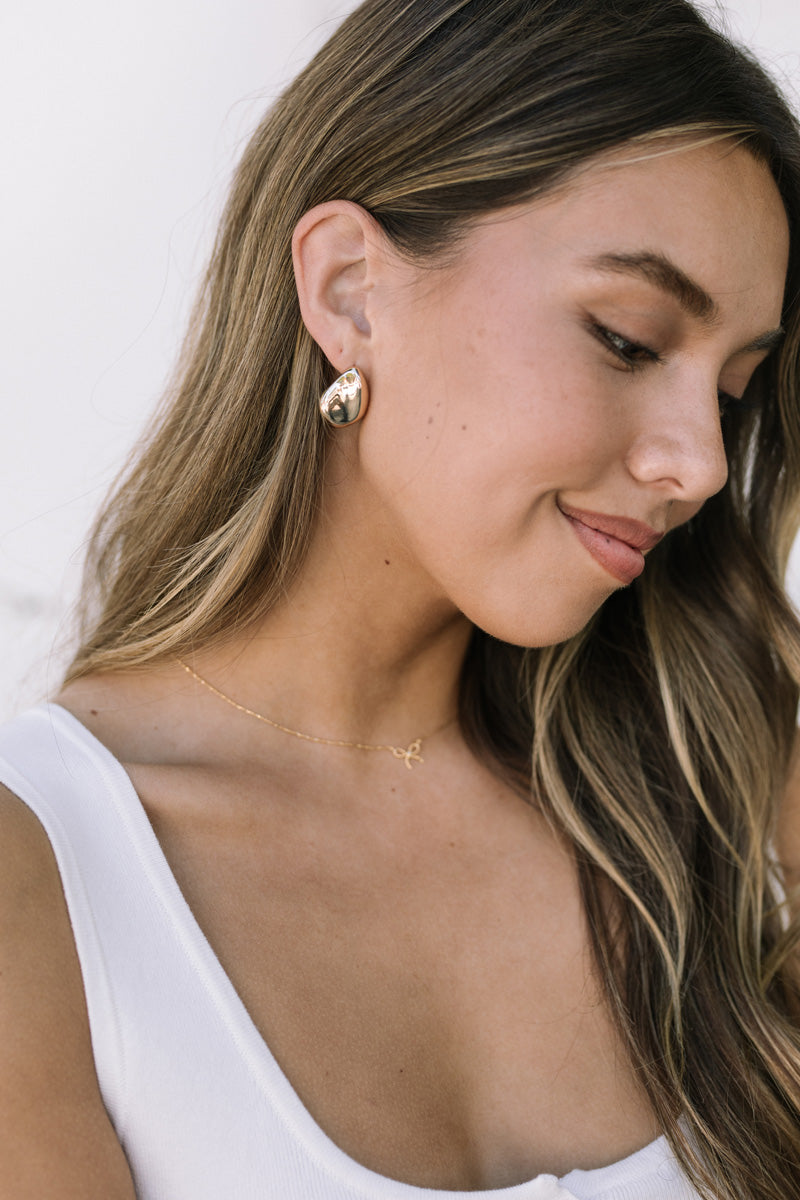 A close-up portrait of a woman with long, wavy brown hair wearing gold teardrop-shaped earrings.