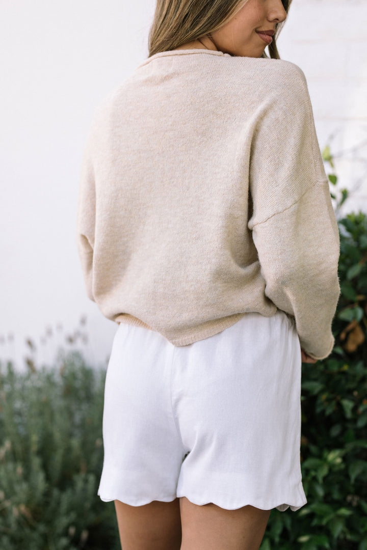 A cozy, beige knit sweater paired with white, scalloped-edge shorts, showcasing a casual yet stylish outfit against a lush, green backdrop.