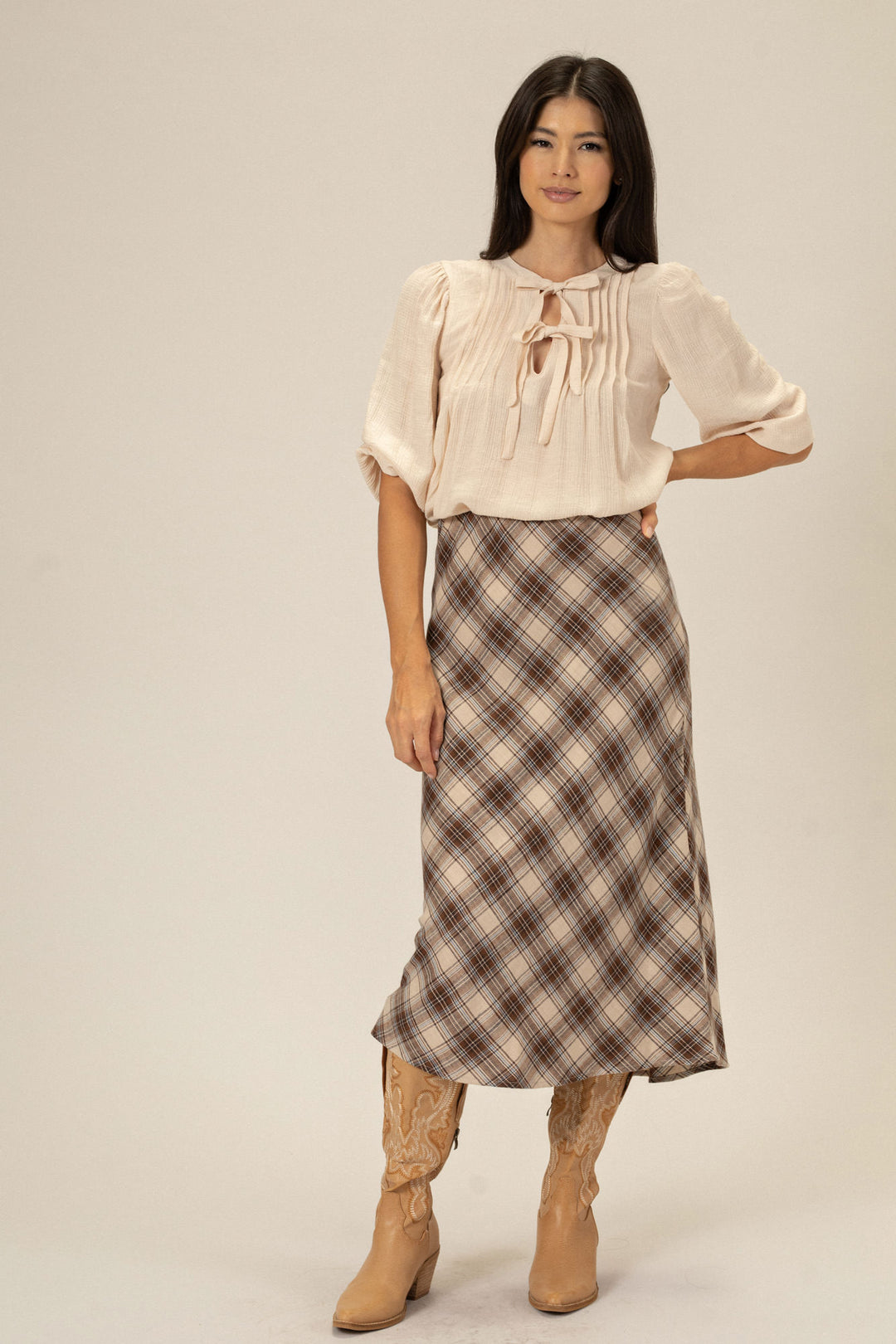 A woman wearing a beige blouse with puff sleeves and a tie detail, paired with a plaid midi skirt in neutral tones and camel-colored cowboy boots.