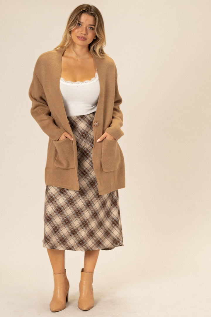 A woman wearing a beige knit cardigan with front pockets, a white lace-trimmed camisole, and a plaid midi skirt, posing in beige heeled boots.