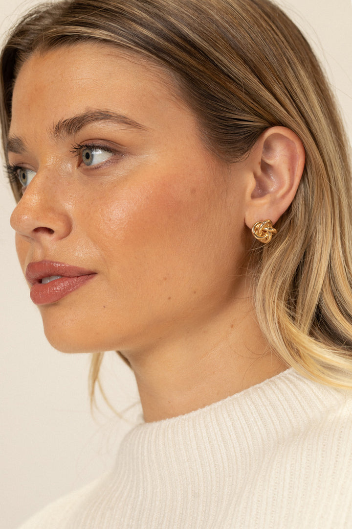 Close-up of a person wearing gold knot-shaped stud earrings, with wavy blonde hair and natural makeup.