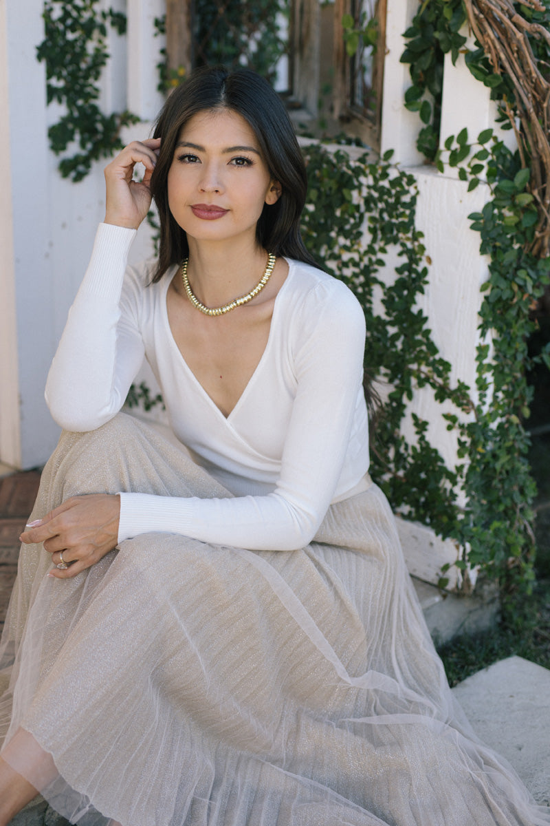 A woman with long dark hair and a white top sits in front of a leafy backdrop, wearing a gold bead necklace.