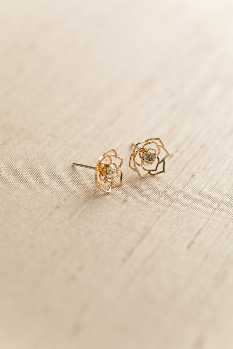 Delicate gold-tone cutout floral earrings with central crystal accents, displayed on a beige textured background.