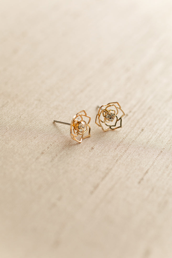 Delicate gold-tone cutout floral earrings with central crystal accents, displayed on a beige textured background.