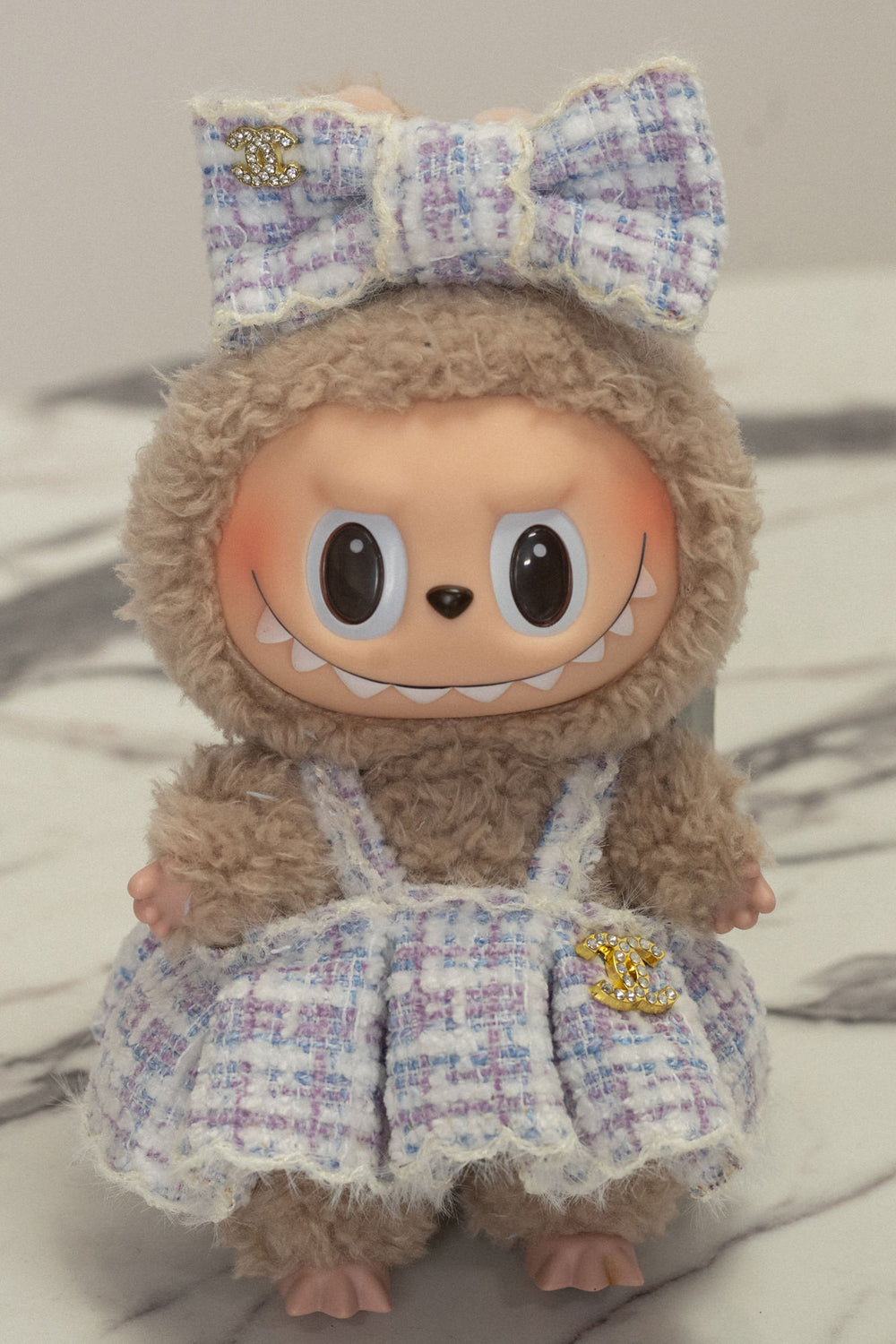 Furry brown plush doll with oversized eyes, wearing a lavender tweed dress with a bow and embellished with gold Chanel-inspired charms.