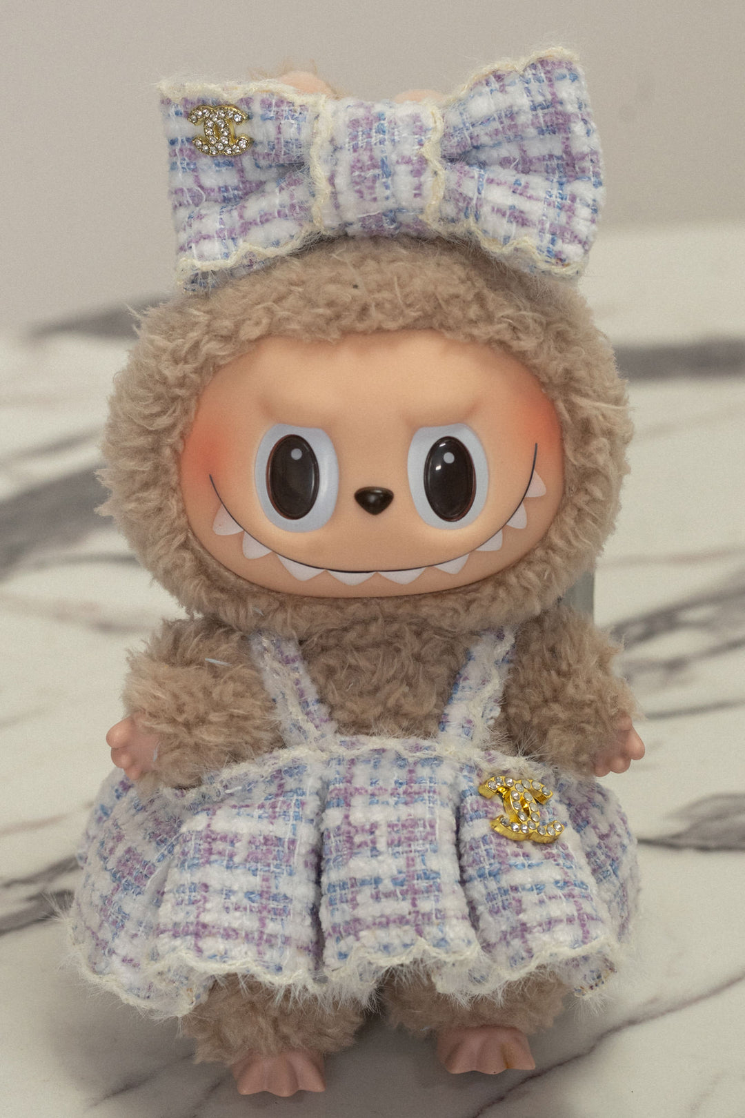 Furry brown plush doll with oversized eyes, wearing a lavender tweed dress with a bow and embellished with gold Chanel-inspired charms.