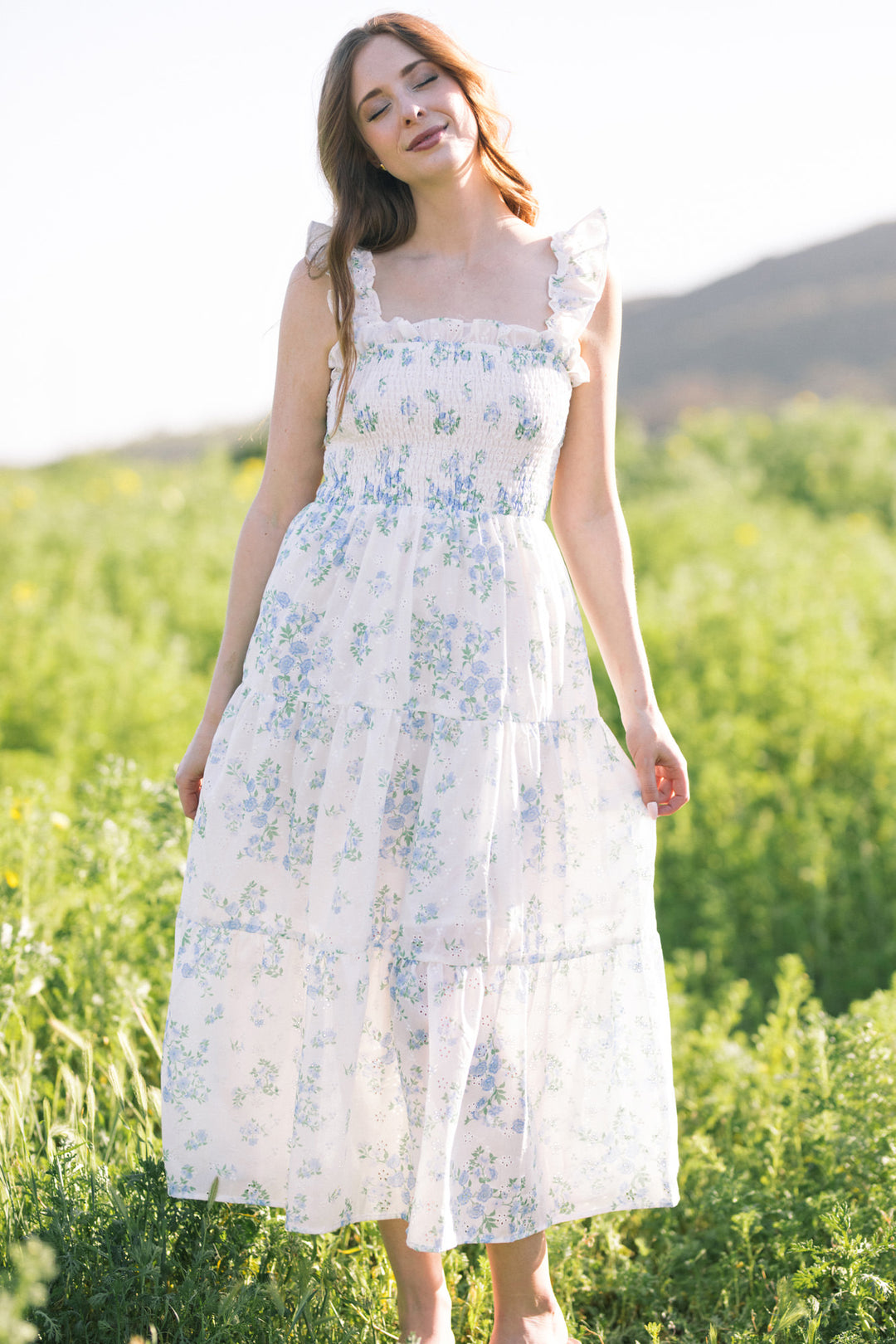 A white floral dress with smocked bodice, ruffled sleeves, and a flowing, ankle-length skirt, worn by a woman with long brown hair standing in a lush green field.