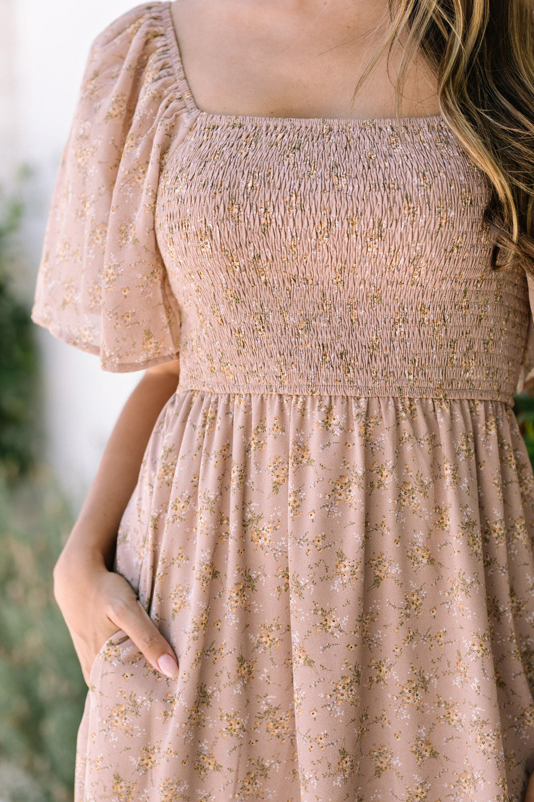 The image shows a pale pink midi dress with a smocked bodice, flutter sleeves, and a floral print throughout the skirt. The dress has a feminine, romantic aesthetic.