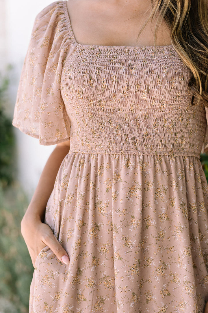 The image shows a pale pink midi dress with a smocked bodice, flutter sleeves, and a floral print throughout the skirt. The dress has a feminine, romantic aesthetic.