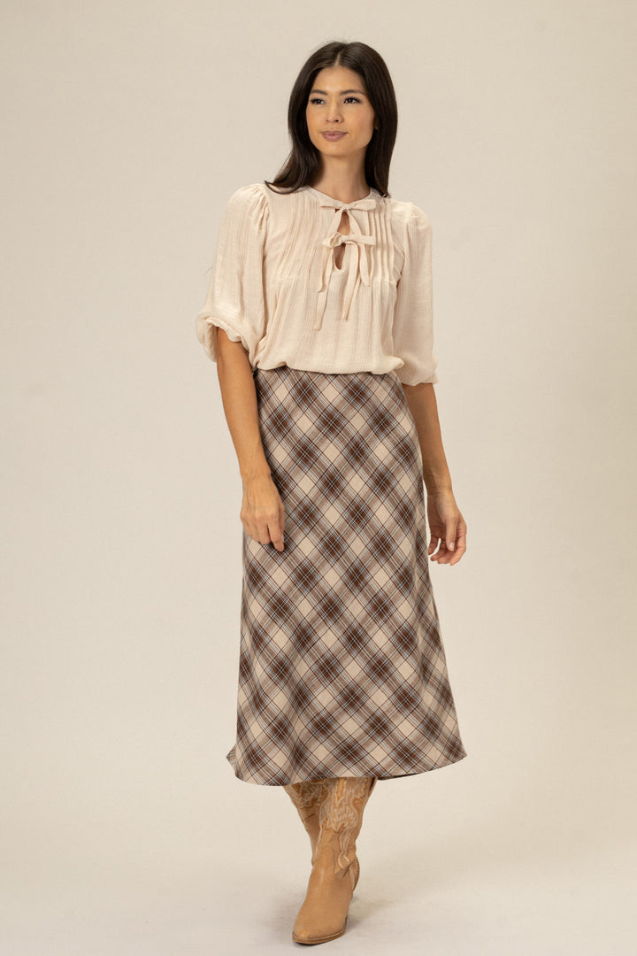 A woman wearing a beige tie-neck blouse and a brown and white plaid midi skirt, standing against a light background.