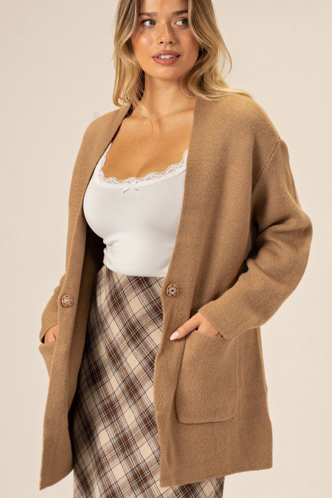 A cozy tan knit cardigan with button closure, worn over a white lace-trimmed tank top and plaid skirt, showcasing the model's casual-chic style.