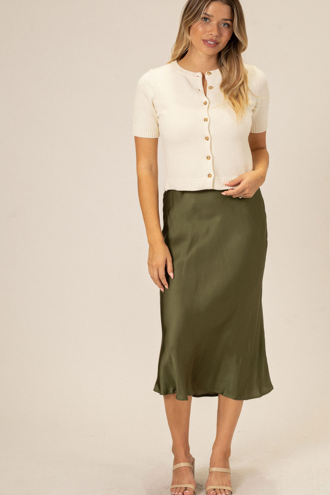 A woman in a white button-up cardigan and olive green midi skirt, standing in front of a neutral background.