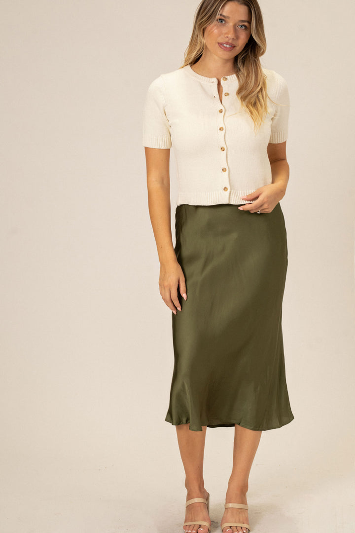 A woman in a white button-up cardigan and olive green midi skirt, standing in front of a neutral background.