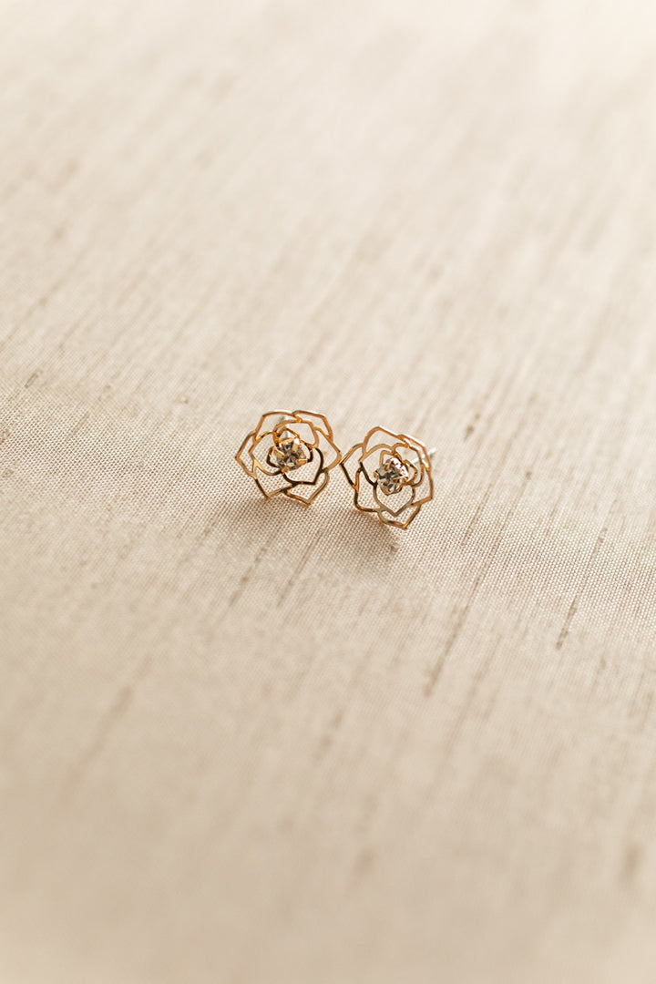 Two delicate rose-shaped gold cutout earrings resting on a textured beige surface.