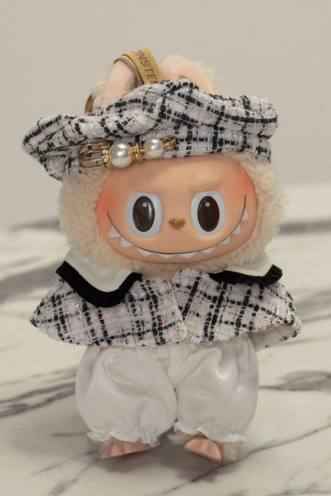 Plush toy with a beige, furry body wearing a black and white tweed beret adorned with pearls and gold accents.