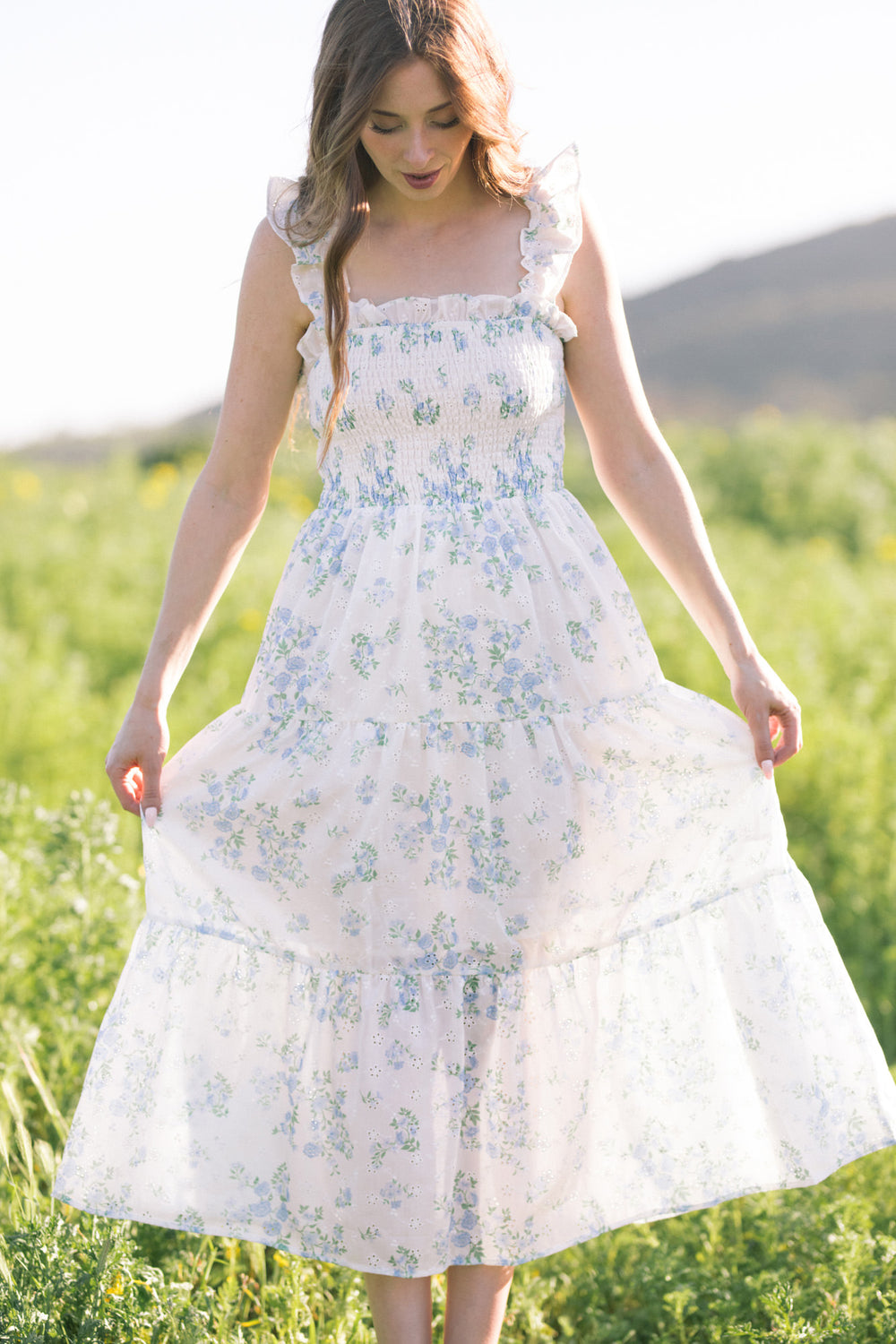 A white floral dress with a smocked bodice, ruffled straps, and a flowing skirt, worn by a young woman with long brown hair in a field of greenery.