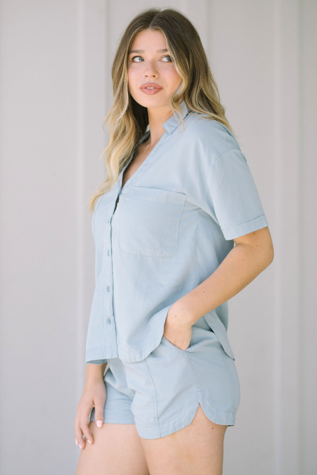 Woman wearing light blue casual button-up shirt and matching shorts, standing with a slight smile.