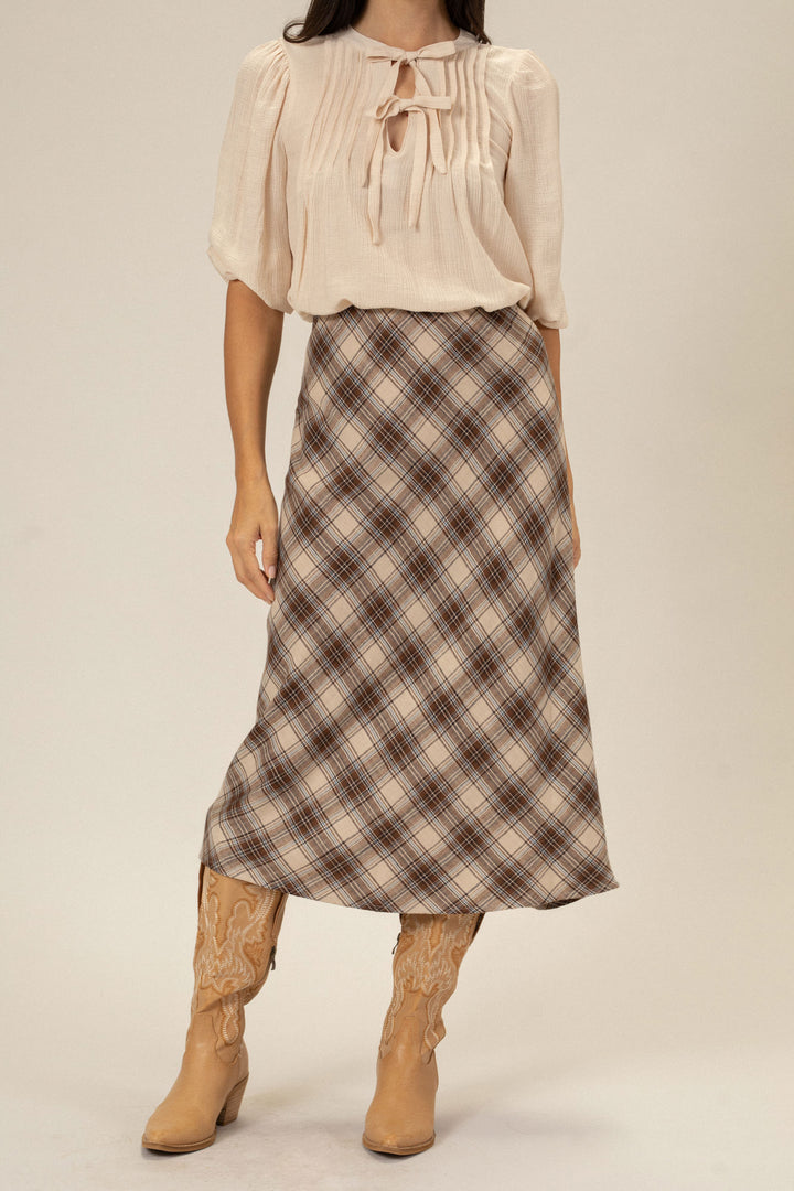 A beige blouse with puff sleeves and a bow tie paired with a brown and beige plaid midi skirt, worn with tan cowboy boots.